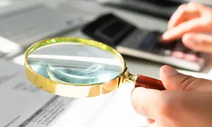 Dłoń trzymająca lupę nad dokumentami, w tle kalkulator i klawiatura.